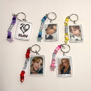 May include: Collection of keychains with clear rectangular frames, each featuring a different image. Some keychains have photos of people, while others have graphic designs. The keychains have beaded accents in various colors and butterfly charms.