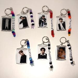May include: A collection of silver keychains with clear rectangular frames, each featuring a photo of a person. Some keychains have beaded accents in various colors, with names spelled out in letter beads. The keychains are for fans of the musical group Monsta X.