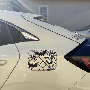 May include: Black vinyl decal featuring bats and spiderwebs, designed for a car's gas tank door. The decal is a Halloween-themed accessory, with a decorative floral design on the car's surface.