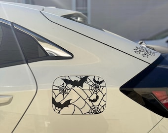 May include: Black vinyl decal featuring bats and spiderwebs, designed for a car's gas tank door. The decal is a Halloween-themed accessory, with a decorative floral design on the car's surface.