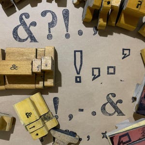May include: A collection of wooden rubber stamps featuring various punctuation marks and symbols including ampersands, exclamation points, question marks, and commas. The stamps are scattered across a surface with black ink impressions of the symbols.
