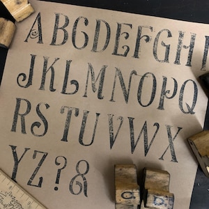 May include: A kraft paper sheet displays a full alphabet printed in black, vintage-style lettering. Wooden letter stamps and a ruler surround the paper. The letters have a distressed, aged appearance.