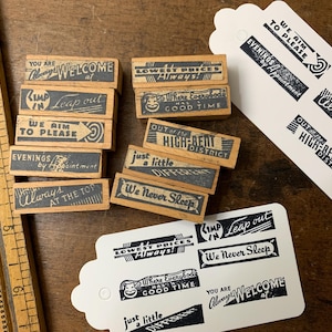 Könnte beinhalten: Eine Sammlung rechteckiger Holzstempel mit schwarzem Text, darunter Phrasen wie We Aim To Please, We Never Sleep und Lowest Prices Always, zusammen mit zwei weißen Papieranhängern mit Stempelabdrücken und einem hölzernen Lineal.