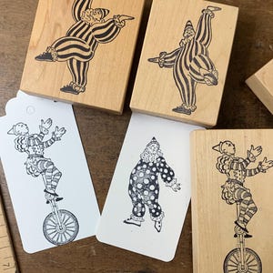 Puede incluir: Tres sellos de goma de madera y dos etiquetas de papel blanco con ilustraciones en tinta negra de payasos. Un sello muestra un payaso en monociclo, otro un payaso con traje a rayas y el tercero un payaso con lunares. Una regla de madera muestra 18 cm.