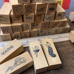 Peut inclure: Une collection de tampons en caoutchouc en bois rectangulaires présentant divers motifs à l'encre noire, dont une ballerine, une personne en robe, le nom Marie, des animaux et des symboles de fête, disposés près d'une règle de 20 cm.