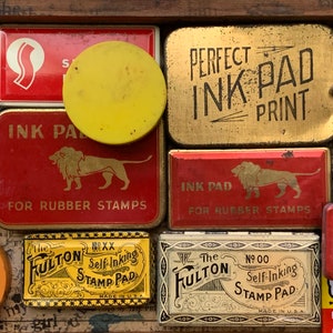 Op de afbeelding: Een verzameling vintage inktkussens en stempelkussens in verschillende vormen en maten. De kussens bevatten tekst als "Perfect Ink Pad Print" en "Ink Pad for Rubber Stamps". Kleuren zijn rood, goud, geel en wit.