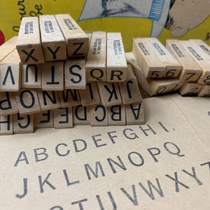 May include: A collection of rectangular wooden rubber stamps featuring black uppercase letters and numbers. The stamps are stacked in a pile, with individual characters visible on the ends. A tan paper sheet displays a printed alphabet and numbers.