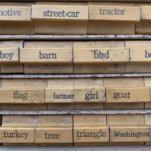 Puede incluir: Primer plano de un juego de bloques de impresión de madera con varias palabras. Los bloques están dispuestos en filas, con palabras como "locomotora", "tranvía", "tractor" y "pájaro" visibles. Los bloques tienen un aspecto vintage y envejecido.