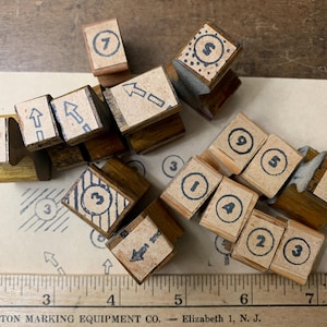 May include: A collection of wooden rubber stamps featuring numbers one through nine, arrows, and geometric patterns, arranged on a surface next to a wooden ruler showing measurements in inches from two to eight, with stamped impressions visible below.