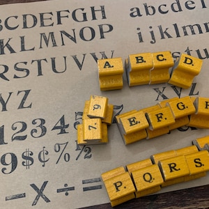 Könnte beinhalten: Eine Sammlung gelber Holzstempel mit Buchstaben und Zahlen, die auf einem bedruckten Papierbogen angeordnet sind. Die Stempel zeigen schwarze Zeichen wie A, B, C, D, E, F, G, H, P, Q, R, S, T und 7. Das Papier zeigt das vollständige Alphabet.