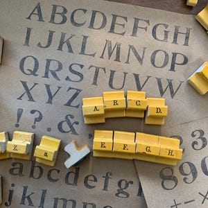 May include: Wooden letter stamps with a yellow base and black lettering are scattered on a brown paper background. The letters A through H are visible, along with other letters, numbers, and symbols. The background features printed uppercase and lowercase alphabets.