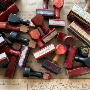 May include: A collection of vintage wooden stamps with red, black, and natural wood handles. Some stamps have text like "Confidential" and "Country File." A ruler is visible at the bottom. The stamps are scattered on a tan surface.