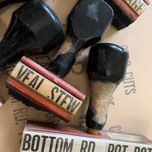 Puede incluir: Cuatro sellos de goma vintage de madera con almohadillas de tinta negra. Los sellos dicen "SAUSAGE", "VEAL STEW", "BOTTOM RD." y "POT RST."