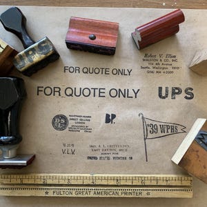 May include: A collection of vintage wooden stamps with black handles and red ink pads, alongside a ruler and text reading "FOR QUOTE ONLY" and "FULTON GREAT AMERICAN PRINTER". The stamps are displayed on a brown paper surface.