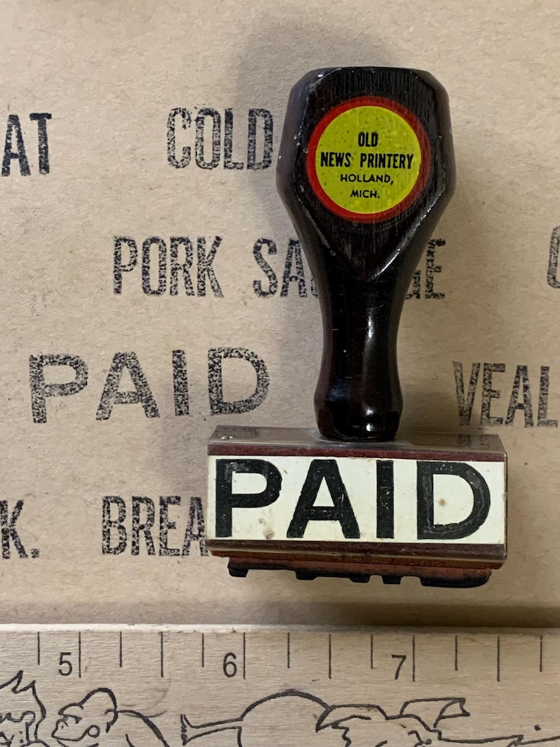 Puede incluir: Un sello de madera con una almohadilla de tinta negra que dice "PAGADO". El sello est&aacute; montado en un mango de madera con una etiqueta circular amarilla y roja que dice "OLD NEWS PRINTERY HOLLAND, MICH."