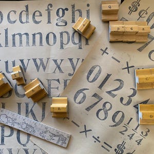 May include: A collection of wooden letter and number stamps with a vintage appearance, arranged on paper printed with letters, numbers, and symbols. A ruler with animal illustrations is also present. The stamps show the letters A, B, C, D, G, H, L, O, P, and Q.