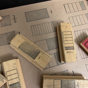 May include: Wooden fraction blocks and fraction diagrams on a tan background. The blocks are rectangular with fraction representations. The diagrams show fractions like 2/5 and 1/6. Some blocks have red ink.