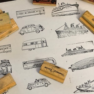 May include: A selection of vintage rubber stamps depicting various modes of transport, such as a bus, car, train, and zeppelin. The stamps are on a white surface.
