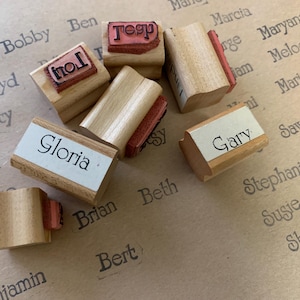 May include: A collection of wooden name stamps, including "Gloria", "Gary", and "Tesp", with red ink pads. The stamps are scattered on a light brown surface with printed names in a cursive font.