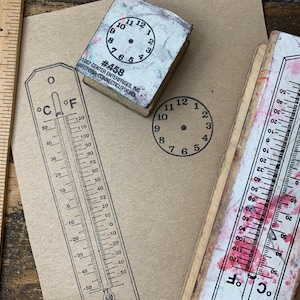 May include: A wooden ruler, thermometer stamps, and a clock stamp are arranged on a tan surface. The thermometer stamps show temperature scales in Fahrenheit and Celsius. The clock stamp displays the numbers 1 through 12.