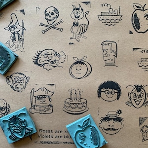 May include: A collection of rubber stamps with various cartoon illustrations, including a skull and crossbones, a devil, a ship, and an apple. Several blue stamp pads are scattered around the images. The stamps are on a tan background.