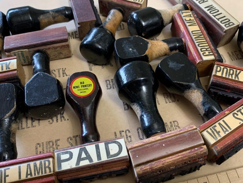 Puede incluir: Una colecci&oacute;n de sellos de goma vintage de madera con almohadillas de tinta negra. Los sellos presentan palabras como "Pagado", "Carne molida", "Redondo de fondo", "Cerdo" y "Bistec de ternera". Un sello tiene una etiqueta amarilla que dice "Old News Printery, Holland, Mich."