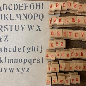 Pode incluir: Um conjunto de carimbos de madeira com letras em tinta vermelha, dispostos ao lado de uma folha de referência impressa mostrando letras maiúsculas e minúsculas. Uma régua de madeira é visível à esquerda, com medidas até 17,78 cm.