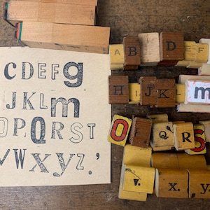 May include: A collection of wooden alphabet stamps and a printed paper sheet displaying the letters A through Z in various fonts and styles. The stamps are arranged in groups, featuring both uppercase and lowercase letters in black, red, and yellow ink.