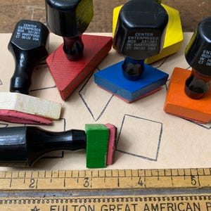 May include: A collection of six wooden stamps with black handles, each marked Center Enterprises Box 331361 W. Hartford CT 06133 USA. The stamps feature various geometric shapes in red, blue, orange, green, yellow, and white, positioned near a wooden ruler.