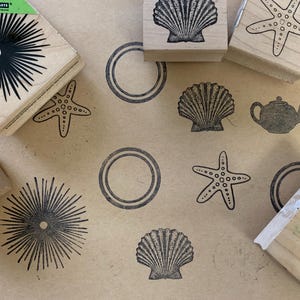 May include: A collection of wooden rubber stamps featuring black ink designs of a sea urchin, a starfish, a seashell, and a teapot, alongside stamped impressions of these shapes and concentric circles on a light brown surface.