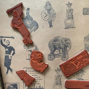 Puede incluir: Una colección de sellos de goma de color terracota con varios diseños de temática circense, incluyendo un artista con una barra de pesas, un elefante sobre ruedas y bloques rectangulares con texto, dispuestos sobre una superficie con ilustraciones vintage.