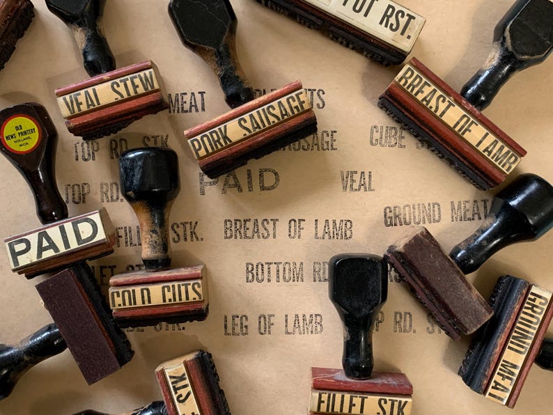 Puede incluir: Una colecci&oacute;n de sellos de madera vintage con varios cortes de carne y etiquetas, incluyendo "Veal Stew", "Pork Sausage", "Breast of Lamb", "Ground Meat", "Paid", y "Top Rd Stk". Los sellos est&aacute;n dispuestos sobre un fondo de papel marr&oacute;n.