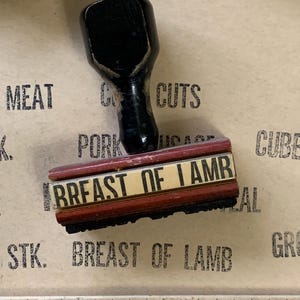 Puede incluir: Un sello de madera rojo y negro con el texto "BREAST OF LAMB" en letras blancas. El sello est&aacute; sobre una superficie marr&oacute;n con otros sellos y una regla.