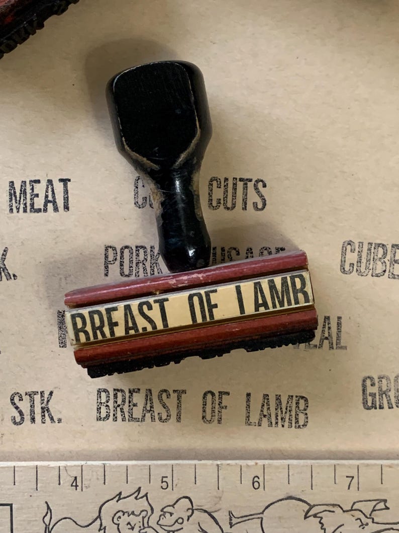 Puede incluir: Un sello de madera rojo y negro con el texto "BREAST OF LAMB" en letras blancas. El sello est&aacute; sobre una superficie marr&oacute;n con otros sellos y una regla.