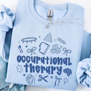 May include: Light blue sweatshirt with the text "Occupational Therapy" in navy blue. The text is surrounded by various blue icons related to occupational therapy, such as a puzzle piece, scissors, a brain, and a lightbulb.