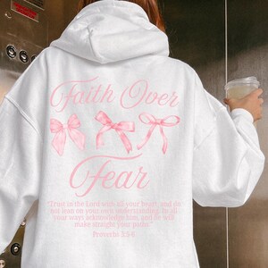 May include: White hoodie with the text "Faith Over Fear" in pink script and three pink bow illustrations. The back of the hoodie also features a Bible verse in a smaller font. The hoodie is a casual, comfortable style.