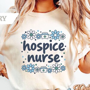 May include: An ivory short-sleeved t-shirt featuring a graphic print with the text hospice nurse in dark blue. The design is surrounded by light blue and teal flowers and small nurse caps with green crosses, arranged in a square pattern.