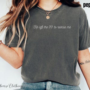 May include: A grey t-shirt with the text "He left the 99 to rescue me" printed on the front. The shirt is being worn by a person with long blonde hair.