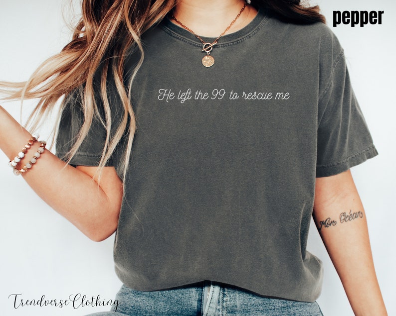 May include: A grey t-shirt with the text "He left the 99 to rescue me" printed on the front. The shirt is being worn by a person with long blonde hair.
