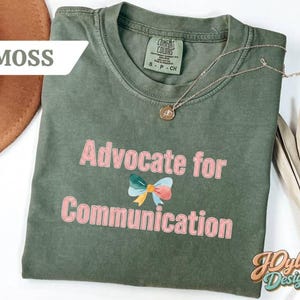 May include: A moss green t-shirt with the text "Advocate for Communication" in pink, featuring a colorful bow graphic. The shirt has a crew neck and short sleeves. A gold necklace is draped across the shirt.