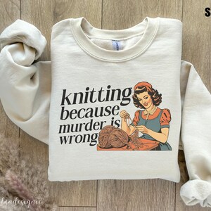 Op de afbeelding: Zandkleurige sweatshirt met de tekst "knitting because murder is wrong" en een vintage illustratie van een breiende vrouw. De vrouw draagt een rode muts en jurk met een blauw schort.