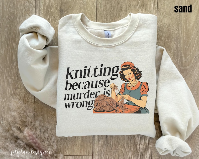 Op de afbeelding: Zandkleurige sweatshirt met de tekst "knitting because murder is wrong" en een vintage illustratie van een breiende vrouw. De vrouw draagt een rode muts en jurk met een blauw schort.
