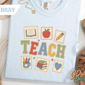 May include: A light blue t-shirt featuring the word TEACH in large, colorful block letters. The design includes illustrations of an open book, a red apple, a pencil, a mug with crayons, a stack of three books, and a heart, all within scalloped borders.