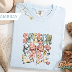 May include: A light blue t-shirt featuring the word SPEECH in large, textured, multi-colored letters decorated with small flowers. Below the text is a large pink bow surrounded by icons including headphones, a microphone, a brain, an ear, a book, a pencil, a puzzle piece, a hand sign, and a heart.