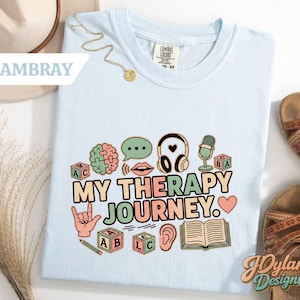 May include: Light blue t-shirt with the text "MY THERAPY JOURNEY" and various graphic icons, including a brain, speech bubble, headphones, microphone, sign language hand, and alphabet blocks. The shirt is a Comfort Colors brand.