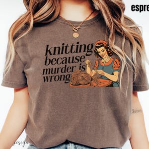 Op de afbeelding: Espresso-kleurig T-shirt met een vintage illustratie van een vrouw die breit en de tekst "knitting because murder is wrong". De afbeelding is in oranje, blauw en bruin. Het shirt heeft korte mouwen.