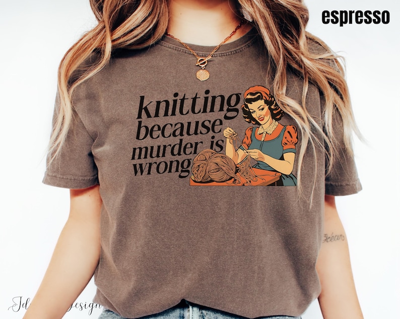 Op de afbeelding: Espresso-kleurig T-shirt met een vintage illustratie van een vrouw die breit en de tekst "knitting because murder is wrong". De afbeelding is in oranje, blauw en bruin. Het shirt heeft korte mouwen.