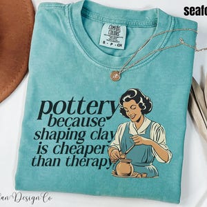 May include: Seafoam green t-shirt with the text "pottery because shaping clay is cheaper than therapy." The shirt features a vintage illustration of a woman working with clay. A gold necklace is draped across the shirt.