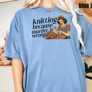 Op de afbeelding: Lichtblauw T-shirt met de tekst "knitting because murder is wrong" en een vintage illustratie van een breiende vrouw. De afbeelding toont een vrouw in een rood topje en blauwe schort, breiend met bruin garen.