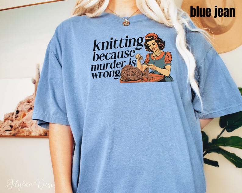 Op de afbeelding: Lichtblauw T-shirt met de tekst "knitting because murder is wrong" en een vintage illustratie van een breiende vrouw. De afbeelding toont een vrouw in een rood topje en blauwe schort, breiend met bruin garen.